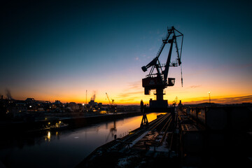 Obraz premium The harbor cranes on the quay wall at sunset. The cranes are silhouetted against the setting sun. 
