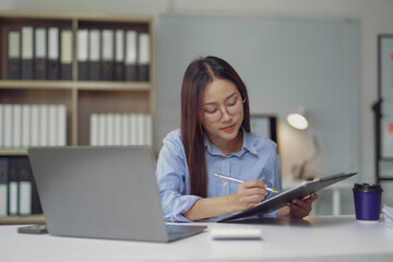Fototapeta premium Asian businesswoman writing notes on clipboard while working at office desk