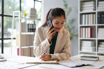 Asian businesswoman talking on phone and taking notes in notebook at office desk