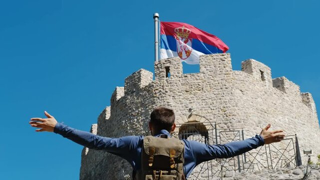 A man stands near the castle with arms raised as if embracing whole world. A popular historical place among tourists. Old Town Fortress in Uzice, Serbia. Castle with Serbian flag on rocky mountain
