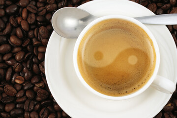 Porcelain cup of coffee with yin-yang shape foam placed on coffee beans, image taken from above.