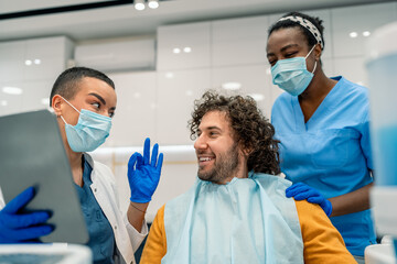 A Caucasian male grins as a female dentist consults with him. A Black nurse assists, both in...
