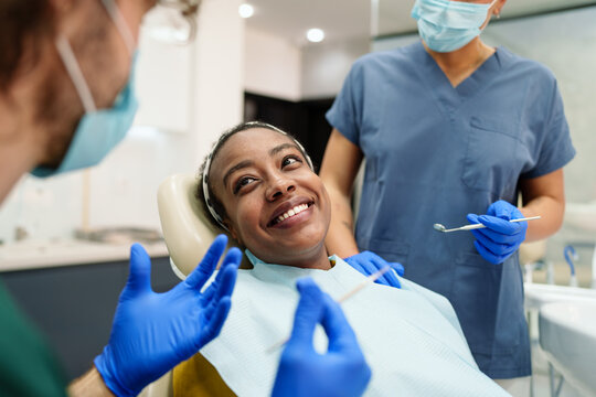 A Black woman smiles warmly while sitting in a dental chair. She is attended by a diverse dental team, including a male and a female, both wearing blue scrubs and gloves. The setting is modern.
