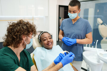 Obraz premium A Caucasian male dentist consults with a Black female patient in a contemporary dental clinic. A female assistant in blue scrubs stands by, ensuring a professional and welcoming environment.