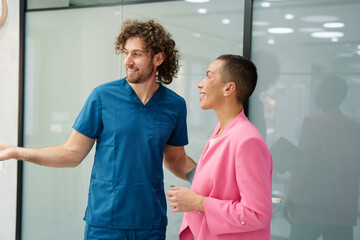 A male Caucasian dentist in blue scrubs warmly greets a female Caucasian patient in a pink blazer within a modern dental clinic. Both are engaged in a friendly conversation, setting a welcoming tone.