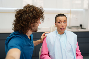Obraz premium A Caucasian male dentist comforts an anxious Caucasian female patient in a modern dental office. She appears nervous while seated in a dental chair, wearing a protective bib.