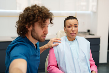 Obraz premium A Caucasian male dentist gently reassures an uncomfortable Caucasian female patient in a modern dental clinic. The dentist wears blue scrubs while the patient dons a pink jacket and dental bib.