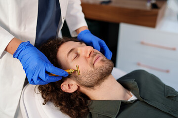 Fototapeta premium A Caucasian male enjoys a rejuvenating facial treatment using a gold beauty bar. A professional beautician in gloves performs the relaxing skincare routine in a modern spa setting.