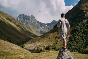 Naklejka premium Man standing on a rock in the Chaukhi mountains, Georgia, looking at the sharp mountain peaks. Green slopes, blue sky, and clouds create a majestic landscape. Concept of travel and freedom