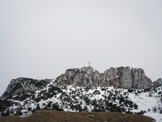 Fototapeta premium A scenic view of the Kampenwand mountain in Bavaria, Germany, featuring a snow-covered landscape, a mountaintop cross, and a small stone chapel under cloudy skies.