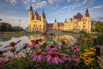 Majestic Moszna Castle on a warm summer morning, surrounded by lush greenery and glowing in soft golden sunlight, reflecting a fairytale-like ambiance perfect for travel and architecture themes.