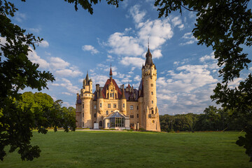 Majestic Moszna Castle on a warm summer morning, surrounded by lush greenery and glowing in soft golden sunlight, reflecting a fairytale-like ambiance perfect for travel and architecture themes.