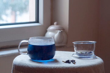 Butterfly pea flower tea in a large sized glass cup on a small table with beige linen tablecloth in front of a window with white tea pot in the background and copyspace