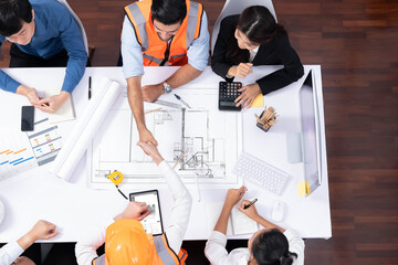 Top panorama of diverse group of civil engineer and client shake hand after make successful agreement on architectural project, review construction plan and building blueprint at meeting table.Prudent