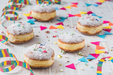 Krapfen, Berliner or donuts with streamers, confetti and mini marshmallows on white wooden background. Colorful carnival or birthday image.