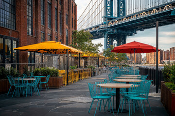 A vibrant outdoor dining area in Brooklyn, New York, offers stunning views of the Manhattan Bridge and the East River. Colorful umbrellas shade the tables and chairs, creating a festive atmosphere.