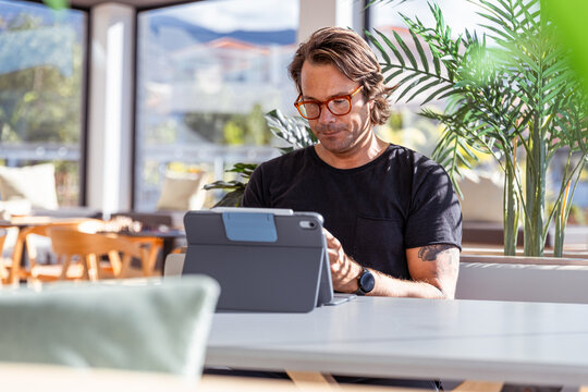 A man is sitting at a table working with his tablet and looking serious and concentrated