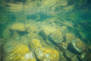Underwater view of a rocky lakebed with crystal-clear water refracting sunlight and a reflection of...