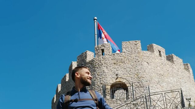 Happy man with backpack stands near the castle. A popular historical place among tourists. Old Town Fortress in Uzice, Serbia. Castle with Serbian flag on rocky mountain