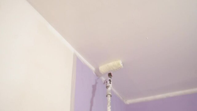 Roller with extension pole applies white paint to ceiling. Interior home improvement. 