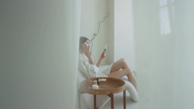 Young woman with under-eye patches looking in small round mirror while sitting in bathroom next to decorative table in morning