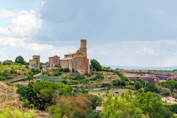 Fototapeta premium Church of San Pietro and two Etruscan towers rising on a hillside in Tuscania, Italy, with lush vegetation, trees, and a cloudy sky creating a picturesque landscape