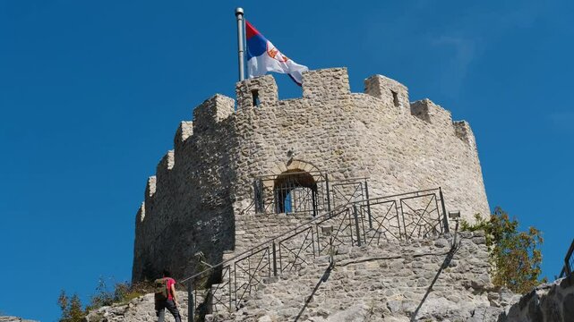 Young woman standing on stone staircase near medieval castle. Old Town Fortress in Uzice, Serbia. Castle with Serbian flag on rocky mountain. Horizontal 4k footage, Travel concept