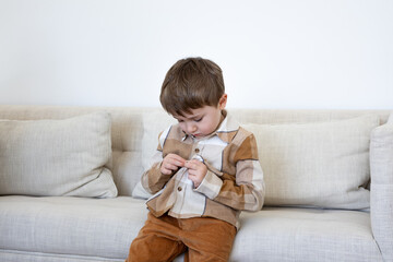 Horizontal view of cute four-year old caucasian boy sitting on beige linen sofa buttoning up his checkered flannel shirt