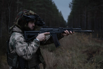 Soldier with night vision device aims assault rifle in forest twilight in Ukrainian combat zone.