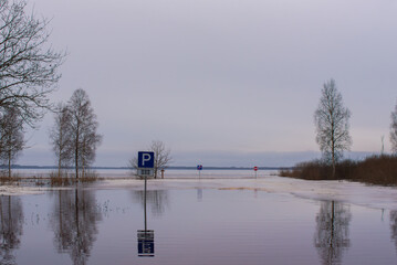  Flooded area with submerged parking signs, trees, and a calm water surface reflecting the overcast sky on a winter day.