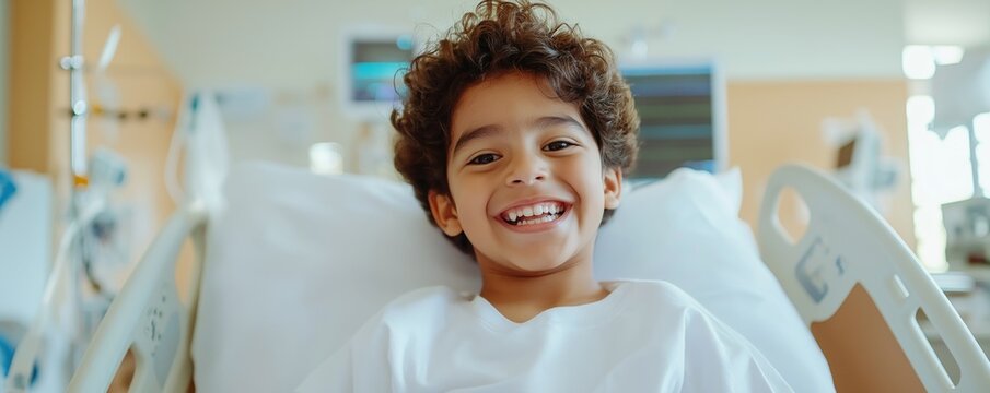 A Hispanic boy around 7 years old sits on his hospital bed with a joyful expression looking at the camera. The bright room is filled with natural light creating an uplifting atmosphere