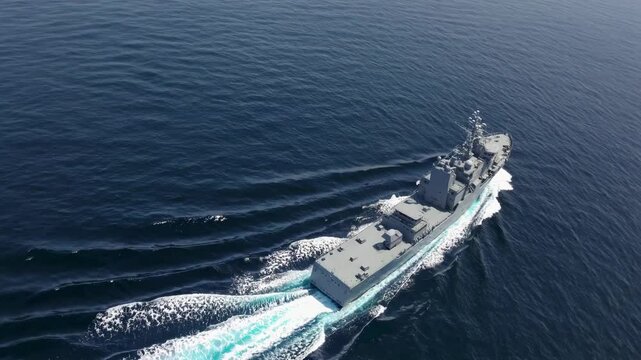 Modern warship cruising through deep blue ocean. Aerial view of naval vessel creating wake patterns. Maritime security and naval power demonstration