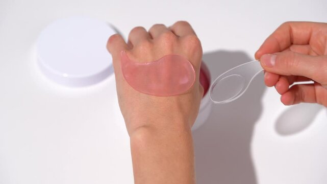 Female hands take out pink patch from jar with spatula and put it on top of her hand on white table background. Concept of beauty products under the eyes for smoothing and moisturizing the skin.