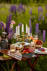 Simple countryside beautiful wedding decor, surprise romantic date outdoors, garden arrangement with lupins flowers on a meadow. Fruits, rose wine, burning candles. Sunset, candlelight, golden hour.