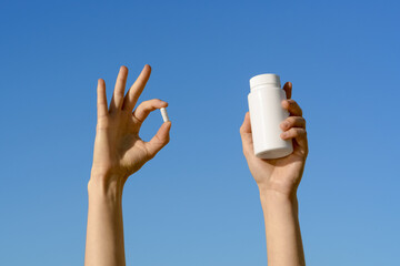 Female hand holding white capsule pill and white mockup jar of pills on blue sky background....