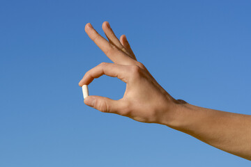 Female hand holding white capsule pill on blue sky background. Concept of pharmacy, medicine, health maintenance and treatment of diseases, dietary supplements and vitamins