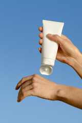 Vertical image of female hands squeezing cream from a white empty tube onto skin against blue sky...