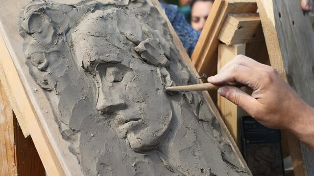 Hand skillfully shapes clay relief on wooden support, capturing delicate features under soft daylight. Neutral tones enhance focus on intricate craftsmanship