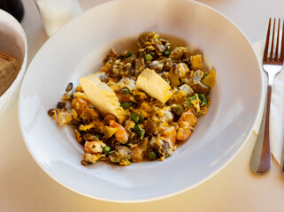 Portion of fried eggs with hearty filling of mushrooms and shrimps are served on white plate. Dish is complemented by two pieces of crispy baguette.
