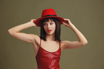 Fototapeta premium Red dress, red hat, woman posing with hands on her head and hair pulled back, looking confident and stylish against a neutral background