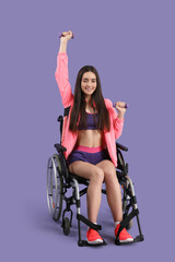 Sporty young woman in wheelchair training with dumbbells on lilac background