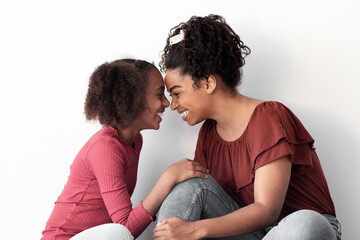 Happy black family cheerful beautiful mother and teen daughter sitting on floor by empty white wall at home, wearing stylish casual outfits, embracing, cuddling, touching with noses, copy space