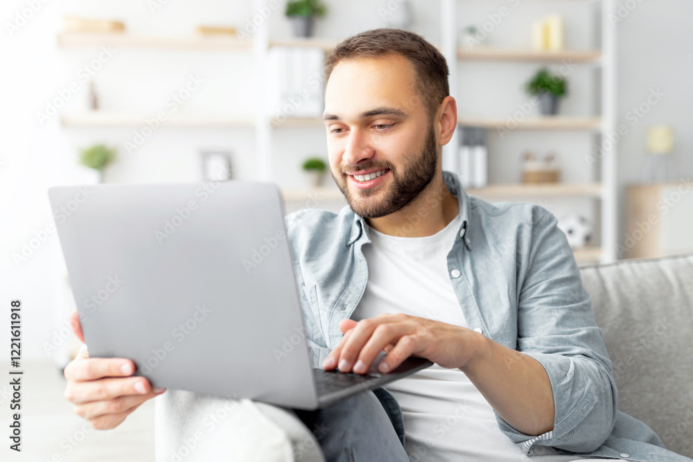 Poster cheerful millennial man sitting on couch with laptop computer, working or studying online from home 
