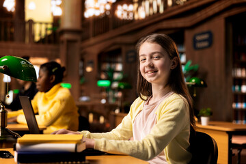 Portrait of adorable teenage girl solving assignments at the school library, feeling ambitious to obtain high grades and finish on a scholarship. Clever scholar smiling at the camera.