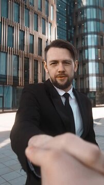4k. POV. Young successful businessman with beard and mustache in suit with documents on textpad walk to the camera, handshake and ask You for sign seriously. Glass building district bg at sunset. Wide