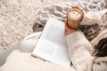 Young woman with blanket, cup of coffee and book sitting on sofa at home, closeup