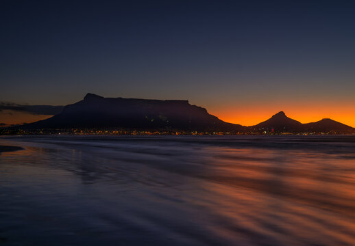 Long exposure shot of Table Mountain silhouette with Cape Town lit up after sunset, viewed from Sunset Beach, South Africa - Powered by Adobe