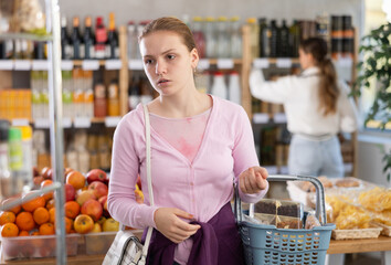 Young woman with grocery basket walking around supermarket and choosing food against the background...