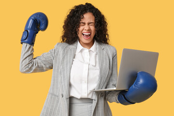 Emotional African-American businesswoman with boxing gloves and laptop on yellow background....