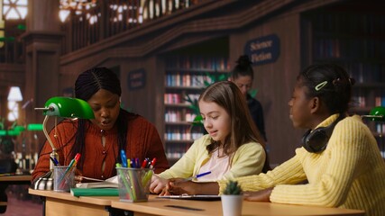 African american private teacher offering support to her students, tutoring them through a lesson plan at a library desk. Tutor providing learning materials, mentoring scholars. Camera B.